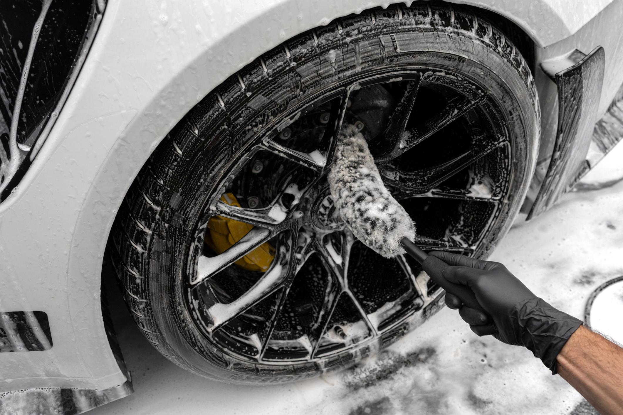 Detailer cleaning a vehicle's alloy wheel with a brush and car shampoo